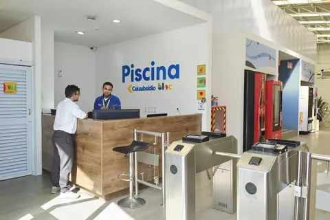 BLOC Piscina Colsubsidio presenta una recepción de madera con empleados y torniquetes de acceso.