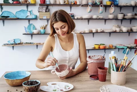 Una joven, con pinceles, pintura acrílica, pinta una taza en los cursos de manualidades Colsubsidio.