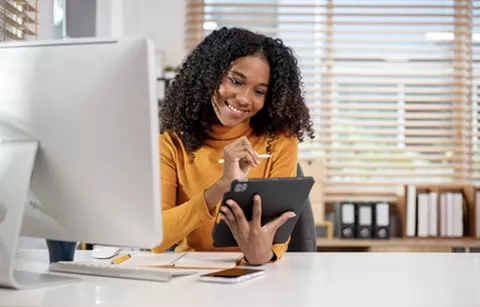 Mujer al frente de su computador, sosteniendo una tablet en la que navega el sitio de la Tienda en Línea de Colsubsidio. 