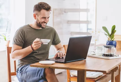 Hombre sonriente sosteniendo una taza de café, al frente de su portátil, conociendo las actividades de la tienda en línea de Colsubsidio.