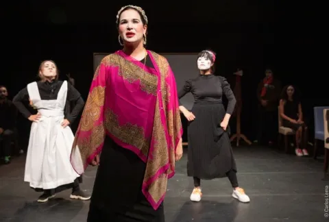 Una mujer con un vestido negro y un chal rosa se para frente a un grupo de mujeres vestidas de negro