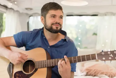 Hombre tocando la guitarra en una caravana con un gato durmiendo en la cama