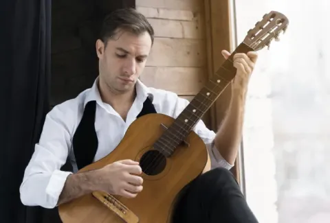 Hombre sentado en el alféizar de una ventana sosteniendo una guitarra