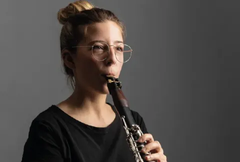 Una mujer con gafas tocando un clarinete sobre un fondo gris
