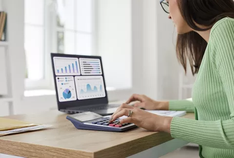 Mujer de suéter verde usa una calculadora junto a un laptop con gráficos financieros, en un escritorio de madera.