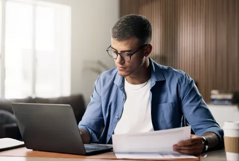Hombre joven de gafas y camisa azul revisa un documento impreso junto a su laptop, en una mesa de madera.