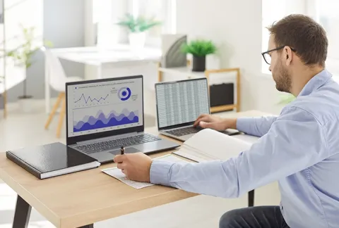 Hombre de camisa celeste trabaja con dos laptops: uno con gráficos financieros y otro con una hoja de cálculo.