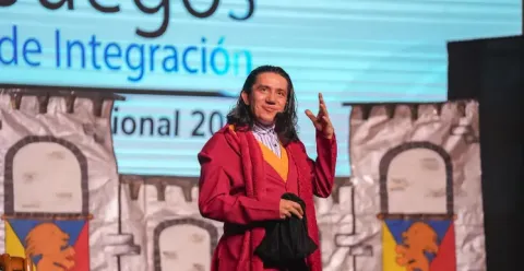 Hombre sonriente en traje rojo y bufanda, gesticulando en el escenario de Teatro Colsubsidio.