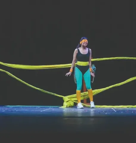Actor en el Teatro Colsubsidio con vestuario vibrante y vendas, rodeado de telas amarillas.