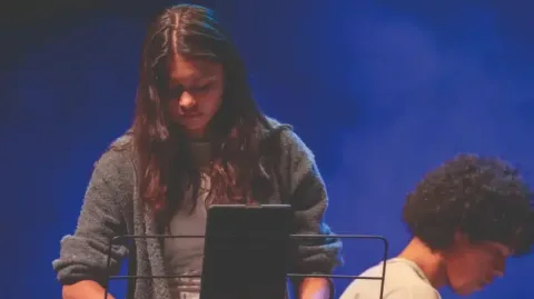 Artista con saxofón en el Teatro Colsubsidio toca frente a micrófono, con chaqueta gris y fondo azul.