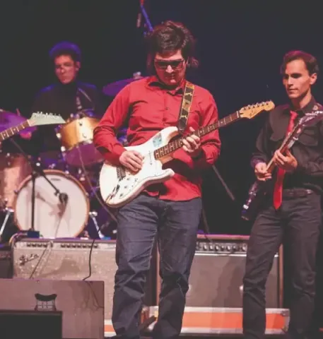 Guitarrista principal de rojo y su banda tocando en vivo en el Teatro Colsubsidio.