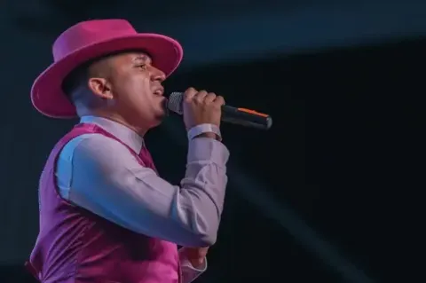 Cantante con sombrero rosa y chaleco canta apasionadamente con micrófono en el Teatro Colsubsidio.