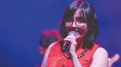Cantante en el Teatro Colsubsidio con blusa roja y collar colorido sostiene micrófono.