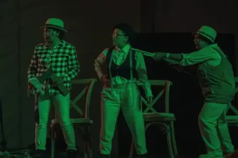 Escena teatral en el Teatro Colsubsidio con tres actores: uno apunta un rifle, otro sujeta un hacha, el tercero mira sorprendido.