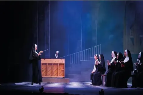 Actuación teatral: una monja dirige un coro, con un pianista, en un escenario de iglesia.