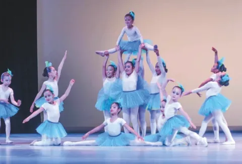 Jóvenes bailarinas de ballet con tutús azules, algunas en una pirámide humana, actúan en el escenario.
