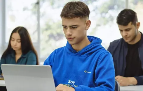 Estudiantes trabajan en laptops y cuadernos, con luz natural y ambiente académico.