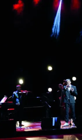 Hombre presentando con micrófono y notas frente a un fondo de sillas luminosas.