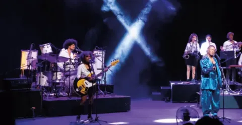 Banda tocando en concierto en el Teatro Colsubsidio con luces azules que forman una X.