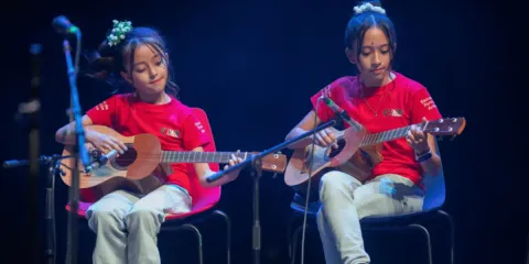Niños en el Teatro Colsubsidio interpretan guitarras pequeñas en escena iluminada.