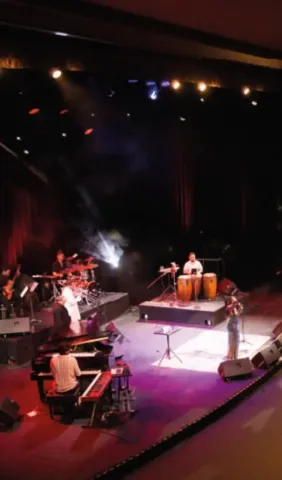 Banda tocando en vivo: músicos con teclado, batería, percusión y vientos en un escenario iluminado.