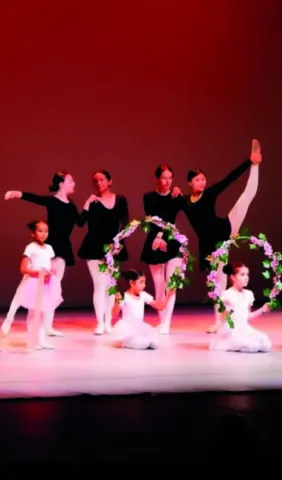 Bailarinas de ballet de diversas edades en un escenario, algunas con coronas de flores.