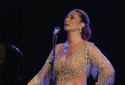 Mujer con un vestido dorado cantando ante un micrófono.