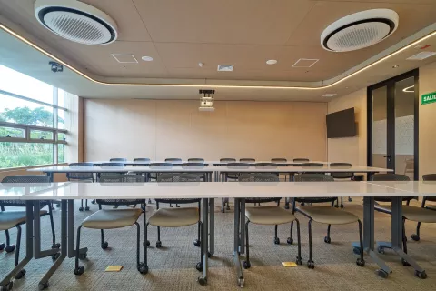 Salón de reuniones modular en Fontanar Colsubsidio con filas de mesas blancas, sillas ergonómicas, videobeam y ventanal.