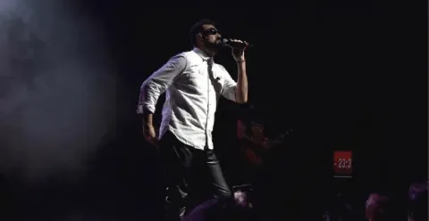 Cantante de rock con camisa blanca y gafas oscuras interpretando una canción en el escenario.