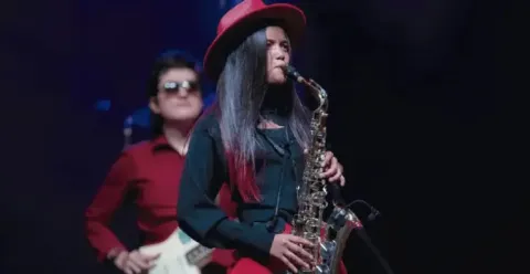 Saxofonista vistiendo sombrero rojo interpretando un solo en el escenario junto a su banda.
