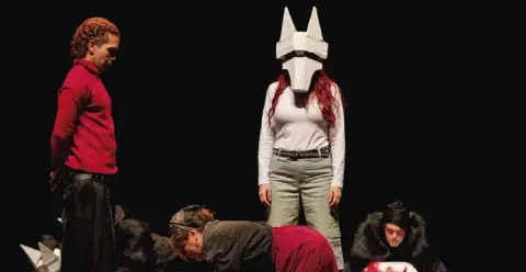 Actores interpretando una escena dramática en el escenario frente a un personaje enmascarado.