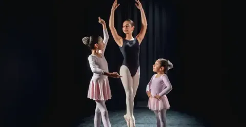 Profesora de ballet realizando una postura en puntas junto a dos niñas estudiantes.
