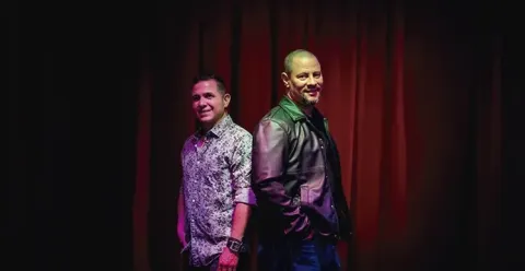 Integrantes del dúo Buena Fe posando sonrientes de espaldas frente a un telón rojo.