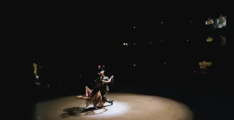 Pareja bailando tango iluminada bajo la luz de un reflector en un escenario oscuro.