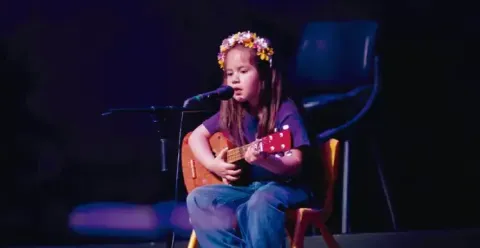 Niña pequeña con corona de flores cantando frente a un micrófono mientras toca un pequeño instrumento de cuerdas.
