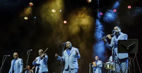 Músicos de la Orquesta Aragón en traje claro interpretando sus instrumentos y cantando bajo las luces del escenario.