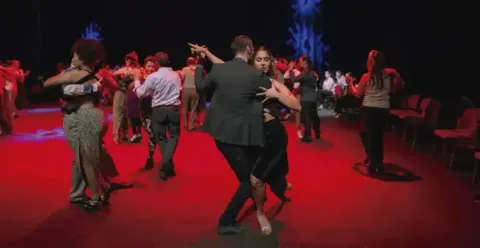 Pareja bailando tango en el centro de una pista rodeada de otros bailarines.