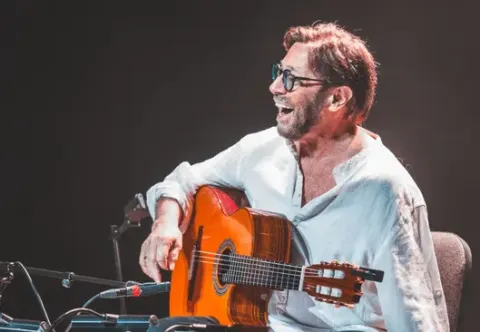 Al Di Meola tocando la guitarra acústica en concierto sobre un escenario iluminado.