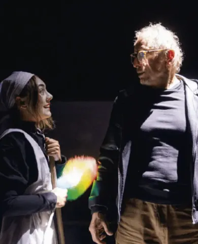 Actriz caracterizada con plumero hablando en el escenario junto a un actor mayor con gafas.