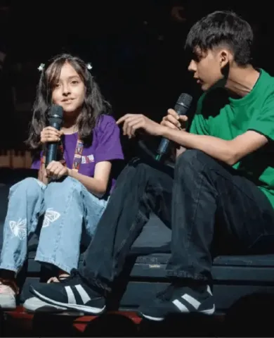 Dos jóvenes sentados en el borde del escenario conversando con micrófonos en mano.