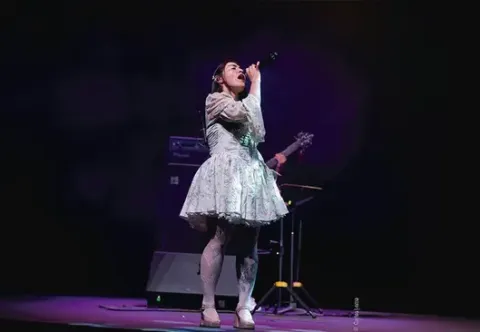 Cantante con vestido floral y mallas en escenario, micrófono en mano, con guitarrista de fondo.