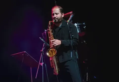 Músico en escenario tocando el saxofón, con micrófonos y batería al fondo.