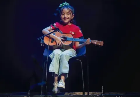 Niña sonriente sentada tocando una guitarra acústica pequeña frente a un micrófono en el escenario.