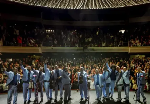 Integrantes de la Orquesta Aragón saludando sonrientes al público desde el escenario de un teatro repleto.