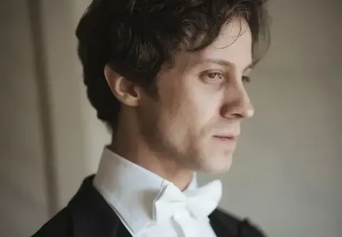 Retrato de perfil del pianista Rafał Blechacz luciendo traje de gala con corbatín blanco.