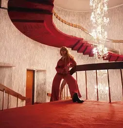Ana Karina Flores vestida de rojo posando cerca a una escalera de caracol e iluminación elegante.