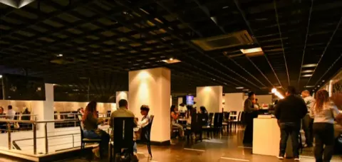 Comensales conversando en las mesas de un amplio café con iluminación cálida y techo reticulado.