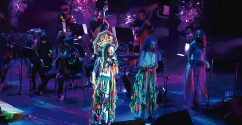 Grupo musical vibrante actuando en un escenario con luces moradas; tres mujeres con trajes coloridos cantan.