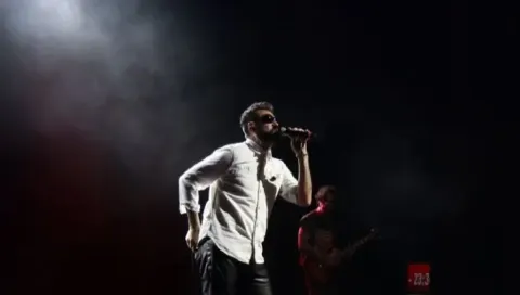 Cantante con barba y gafas de sol en escenario oscuro, micrófono en mano, guitarrista al fondo.