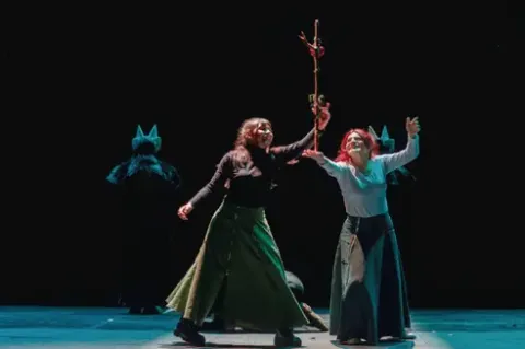 Dos actrices interpretando una escena en el escenario sosteniendo una vara decorada.